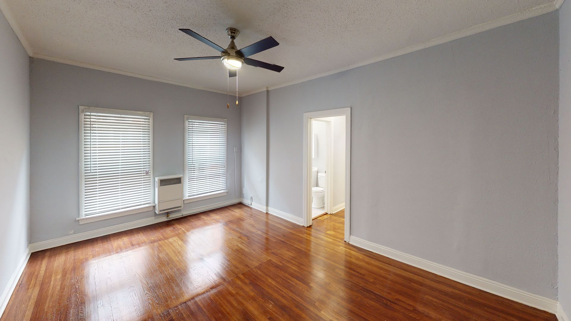 Empty room with wood floors, two windows with blinds, light blue walls, ceiling fan, and a doorway.