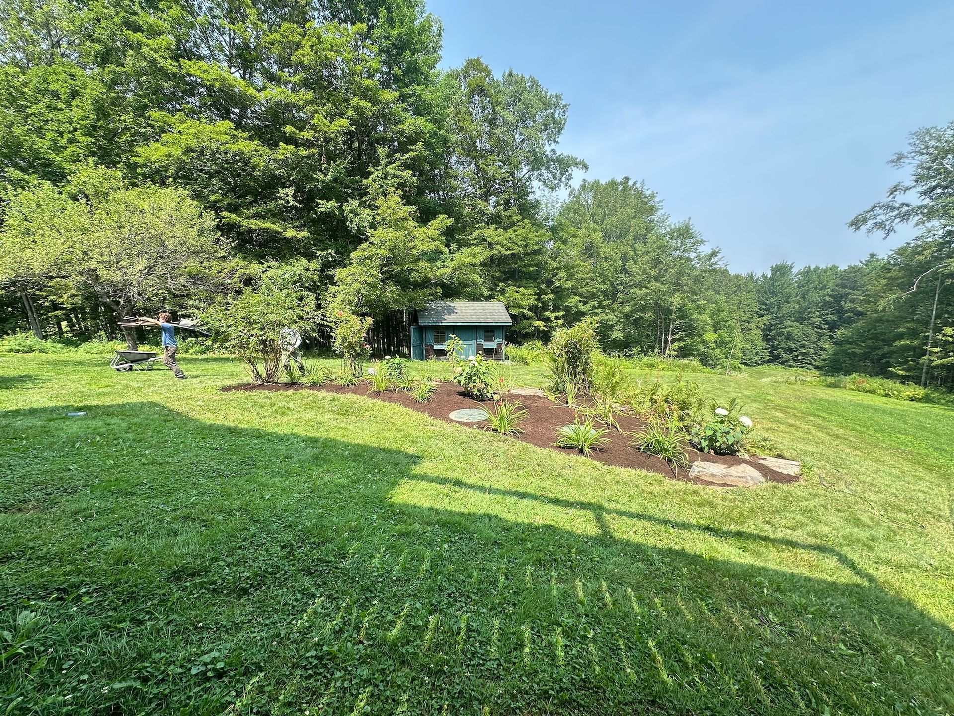 Lush green park with a small garden bed, trees, and a gazebo under a clear blue sky.