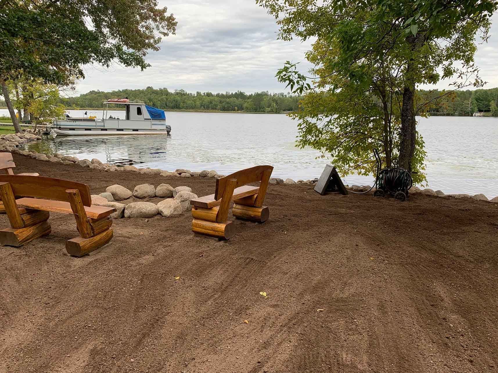 Lakeside fire pit area with log benches, gravel ground, lake view, dock, and overcast sky