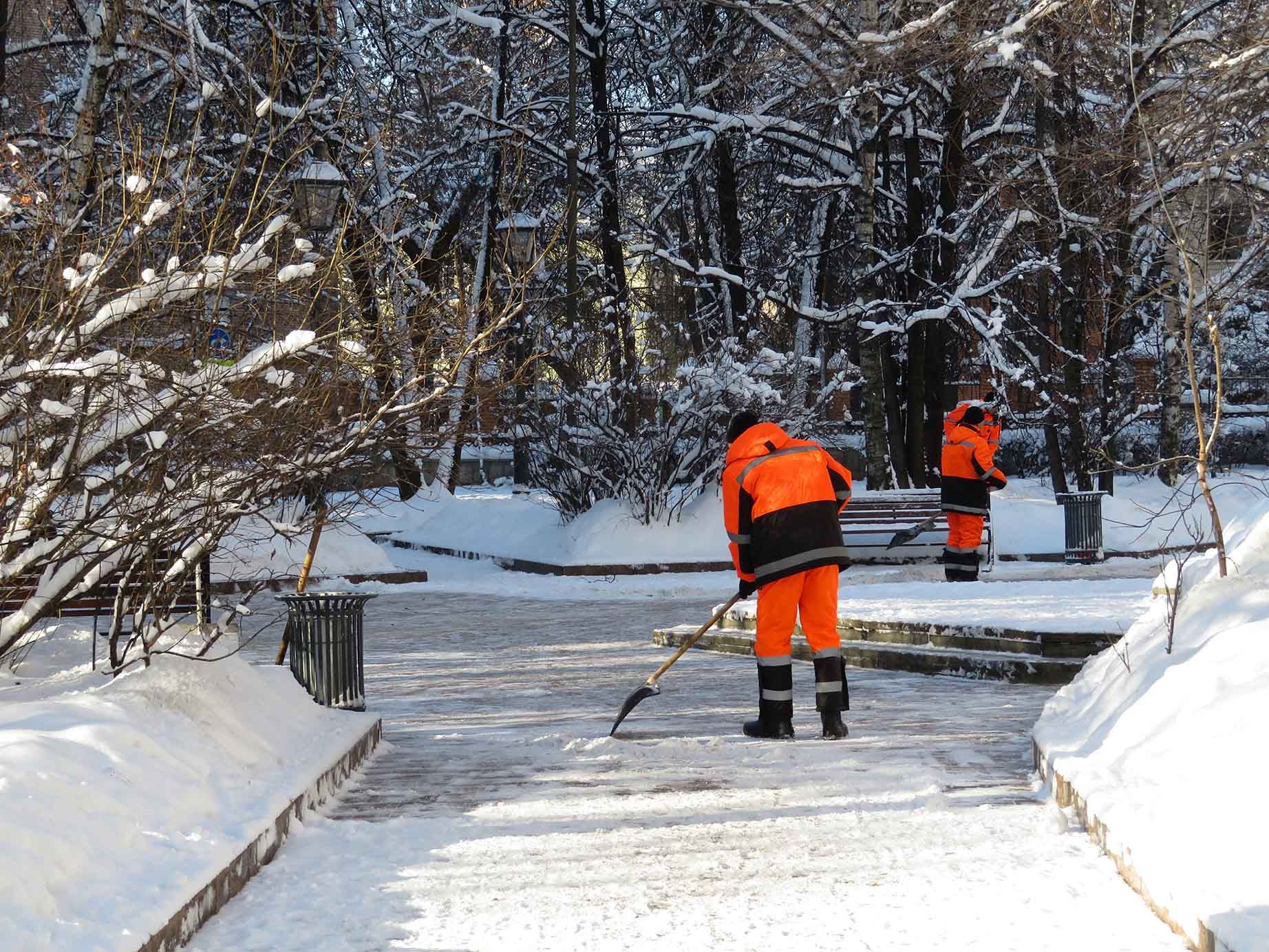 Two workers in orange suits are shoveling snow from a walkway in a park