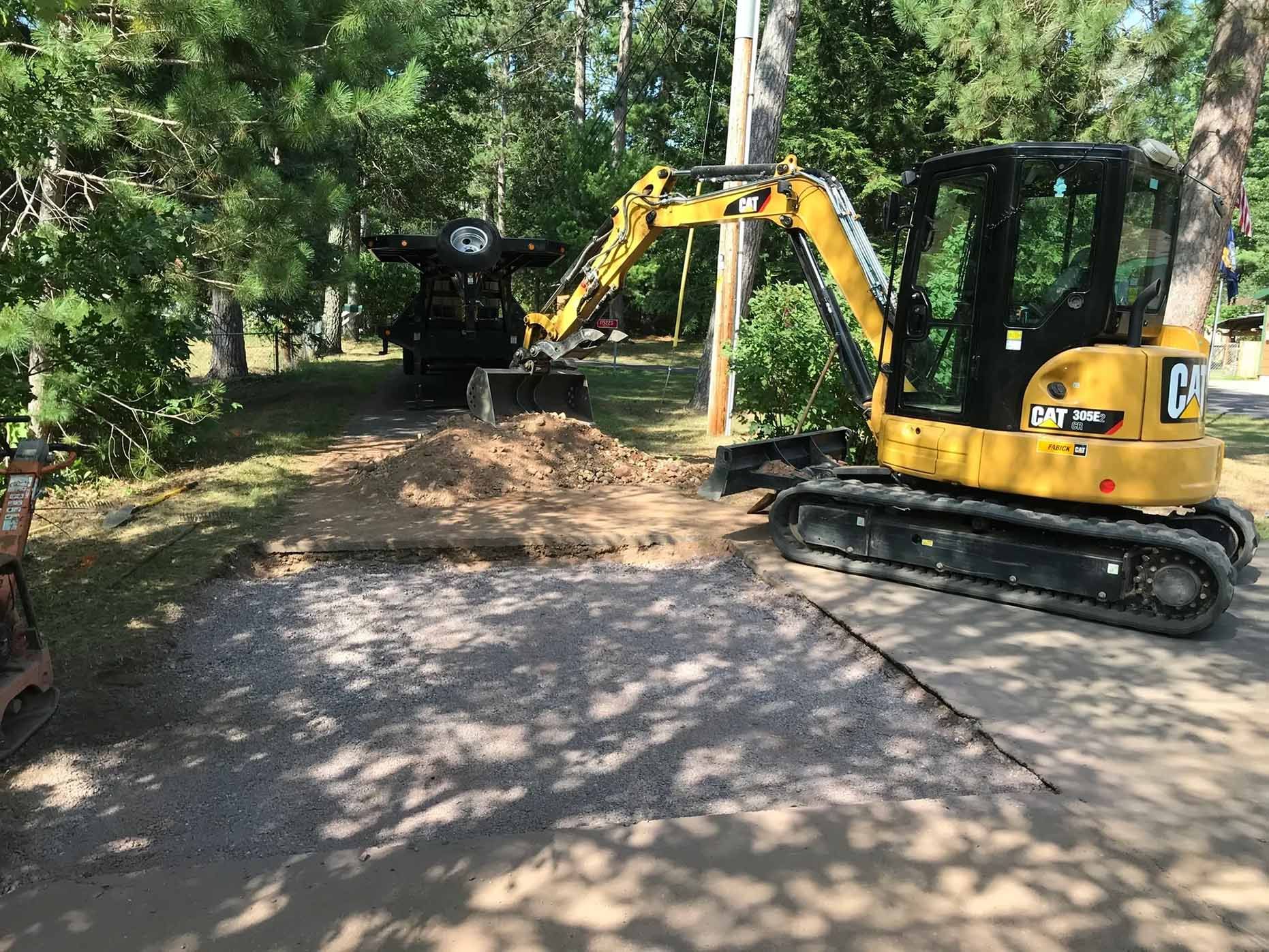 A yellow mini excavator is digging on a gravel driveway with a pile of dirt