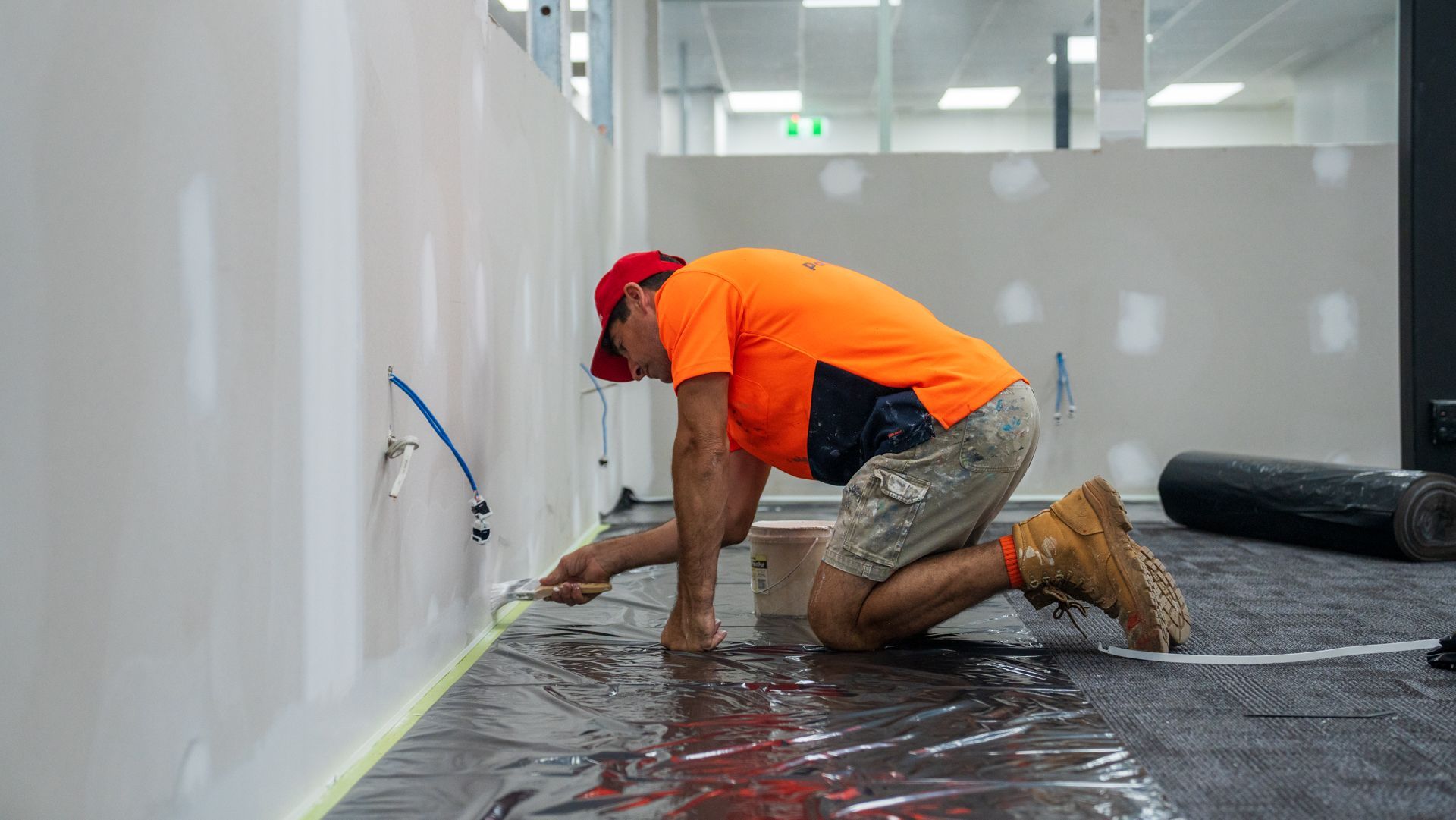 A man is kneeling on the floor painting a wall.