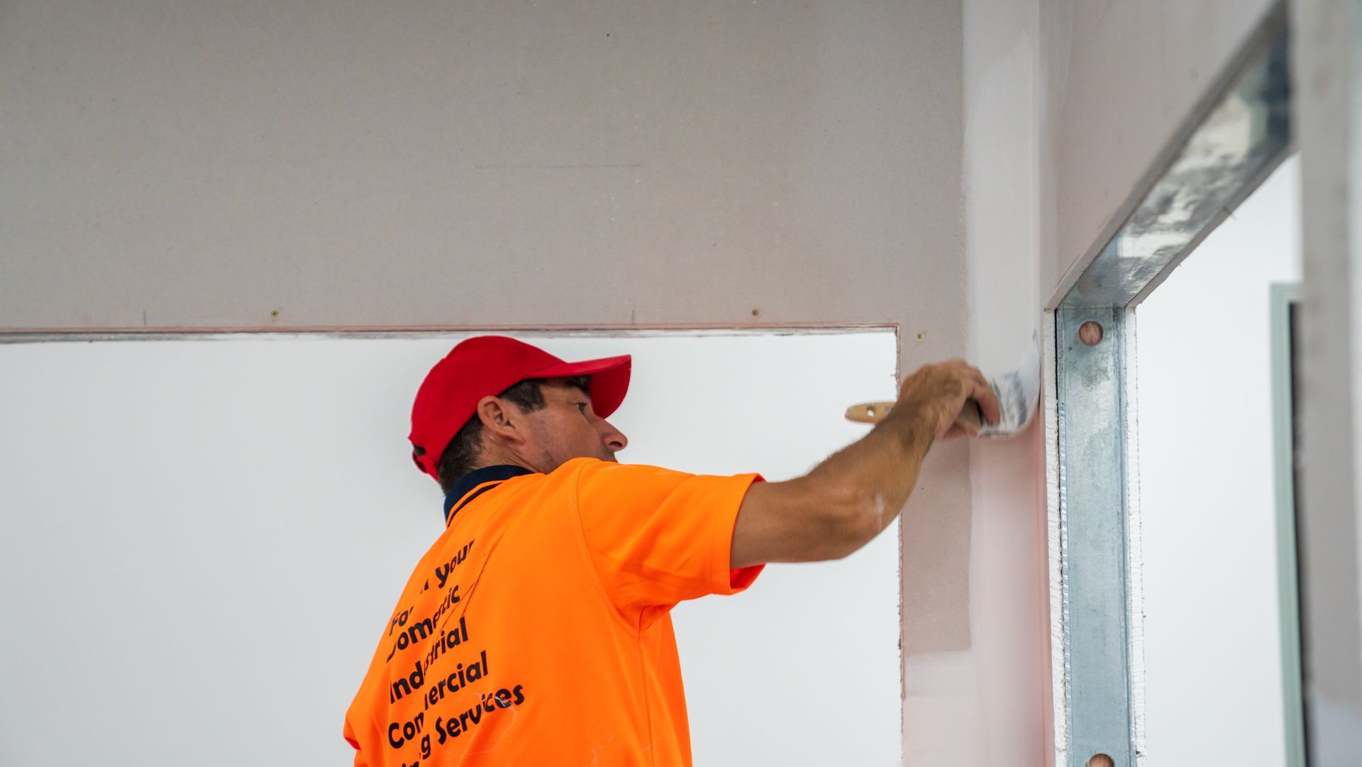 A man in an orange shirt and red hat is painting a wall.