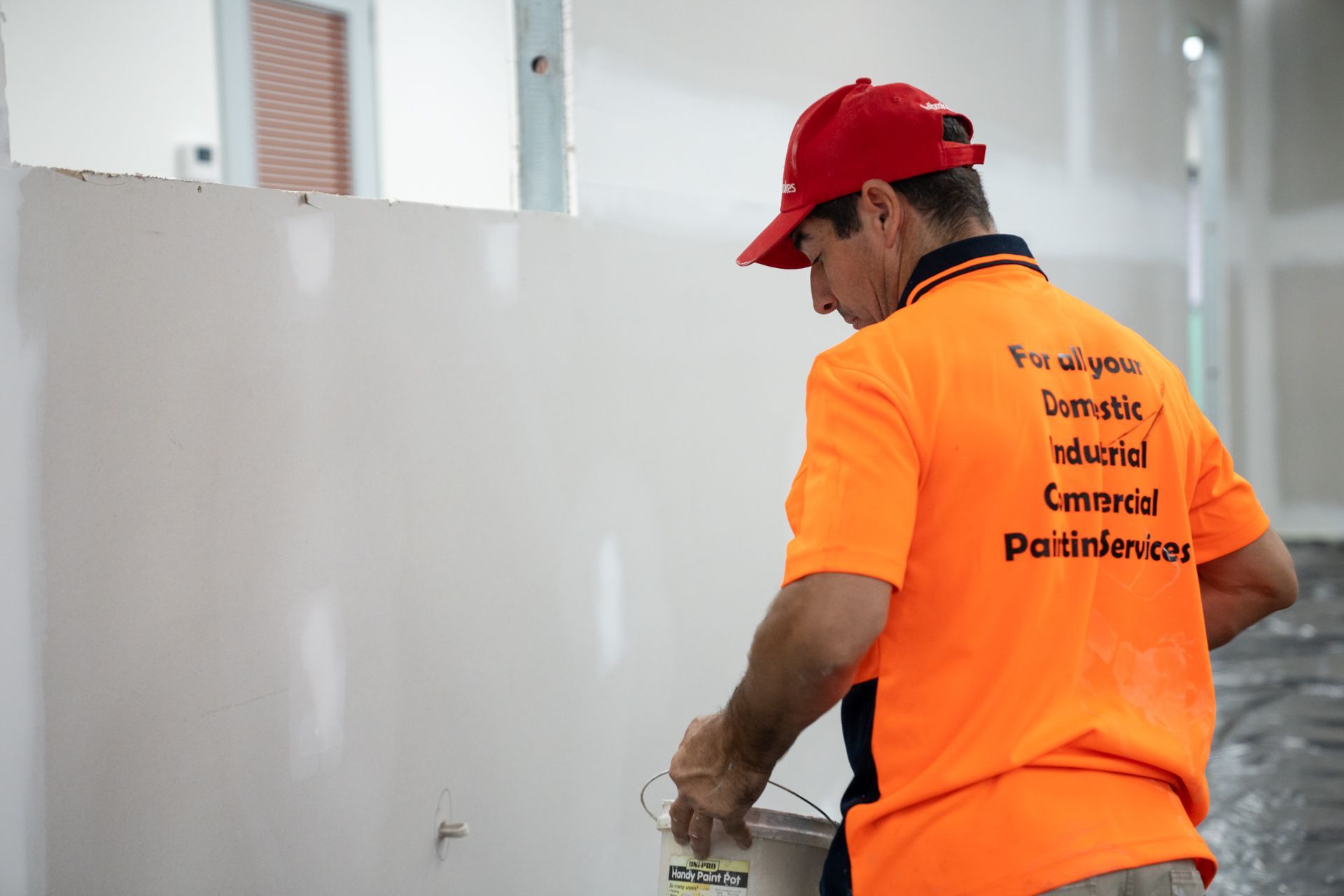 A man wearing an orange shirt and a red hat is painting a wall.