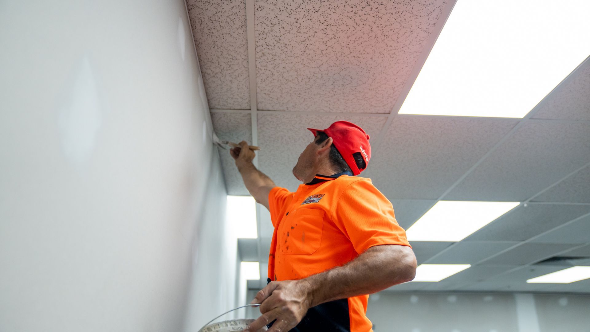 A man in an orange shirt and red hat is working on a ceiling.