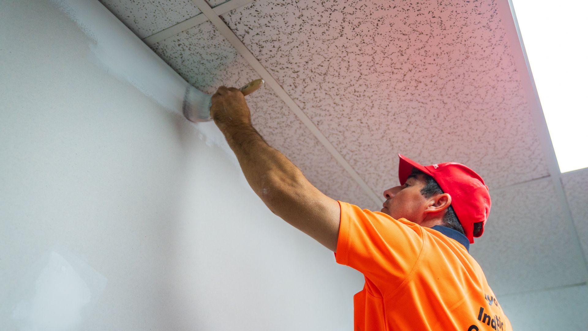 A man in an orange shirt and red hat is painting a wall.
