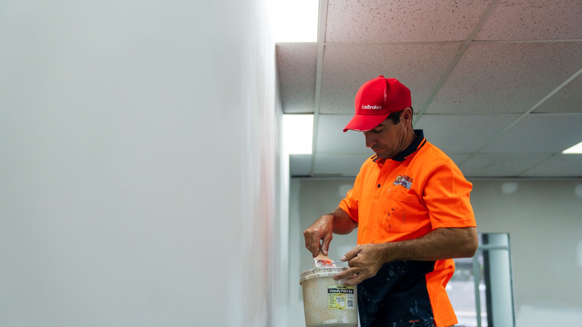 A man in an orange shirt and red hat is painting a wall.
