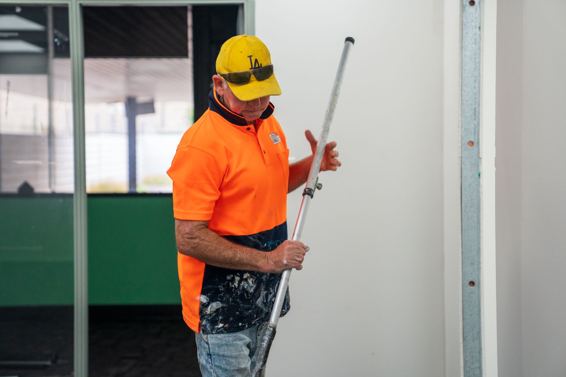 A man in an orange shirt and yellow hat is holding a paint roller.