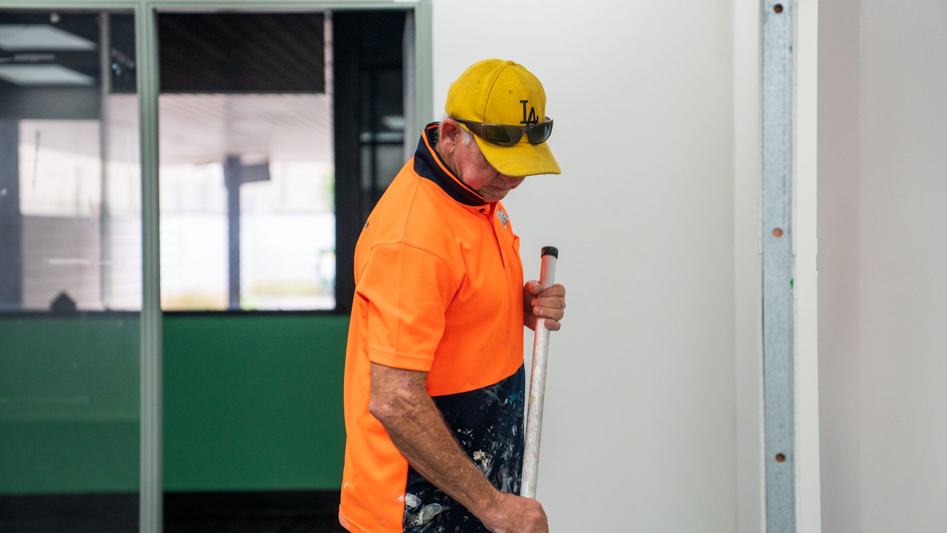 A man in an orange shirt and yellow hat is painting a wall.
