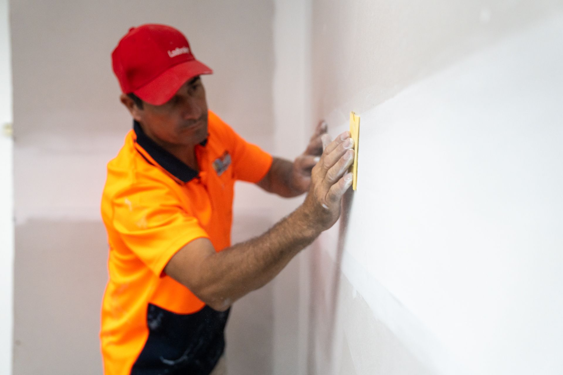 A man in an orange shirt and red hat is sanding a wall.