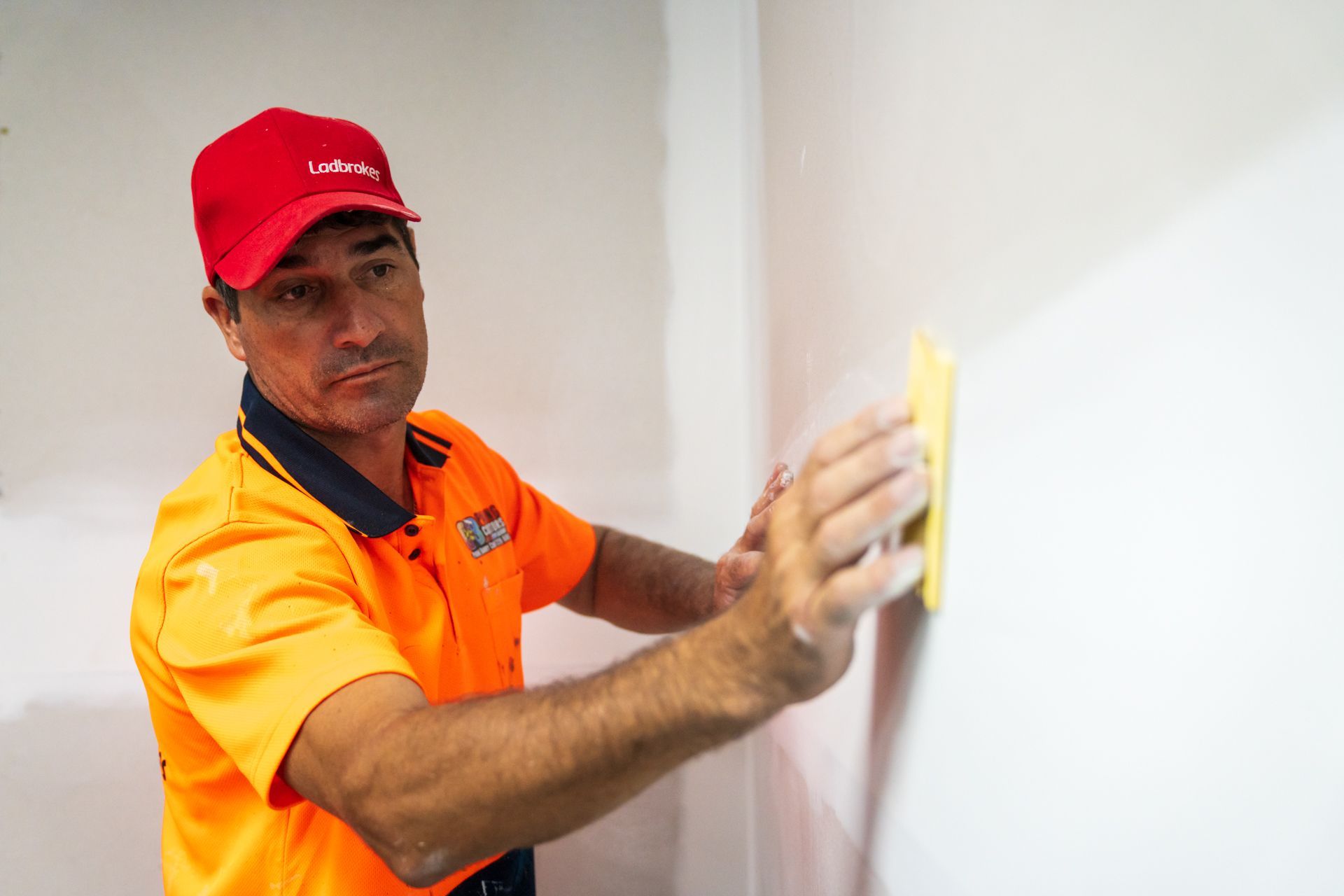 A man in an orange shirt and red hat is cleaning a wall with a sponge.
