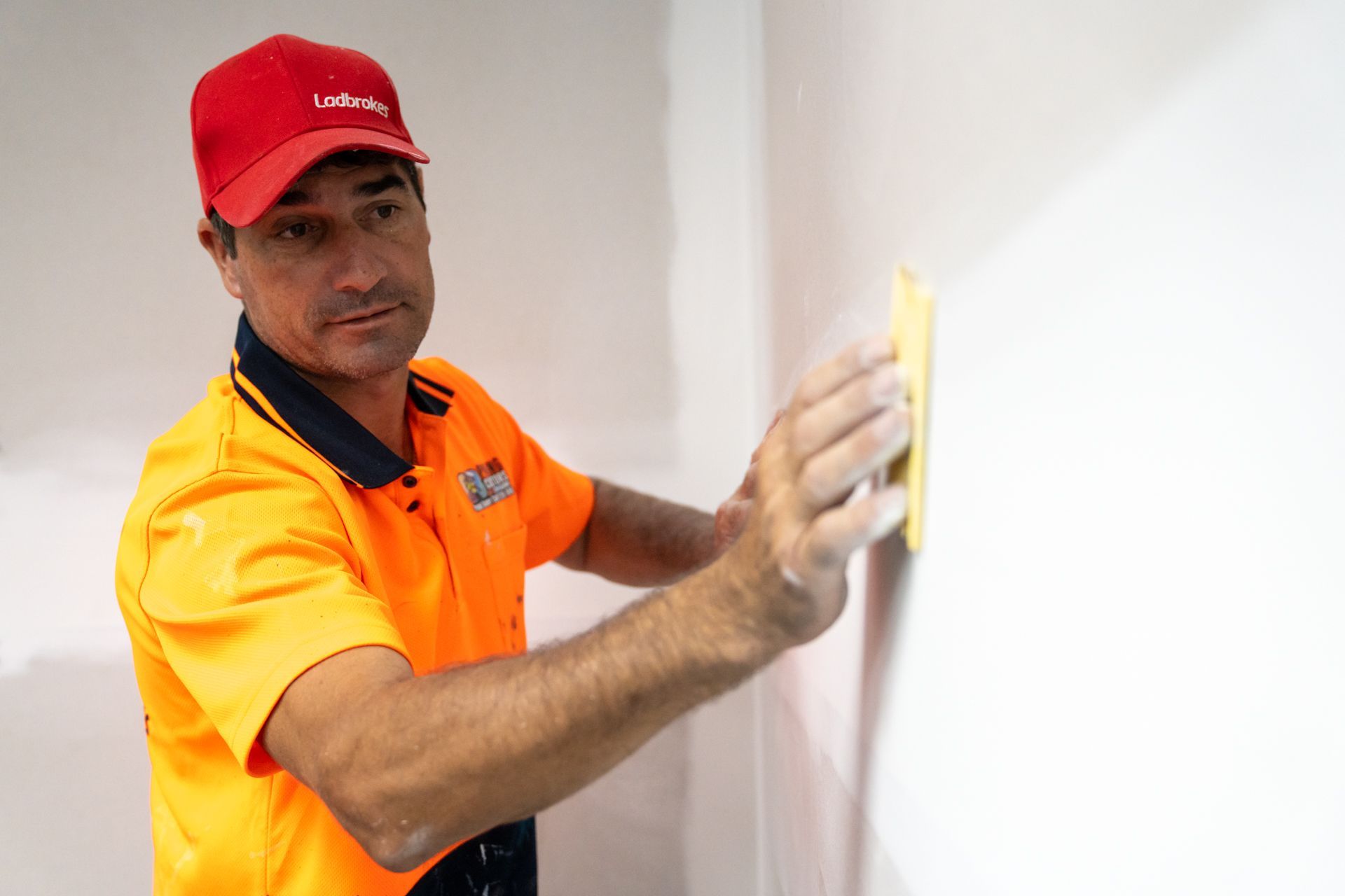 A man in an orange shirt and red hat is plastering a wall.