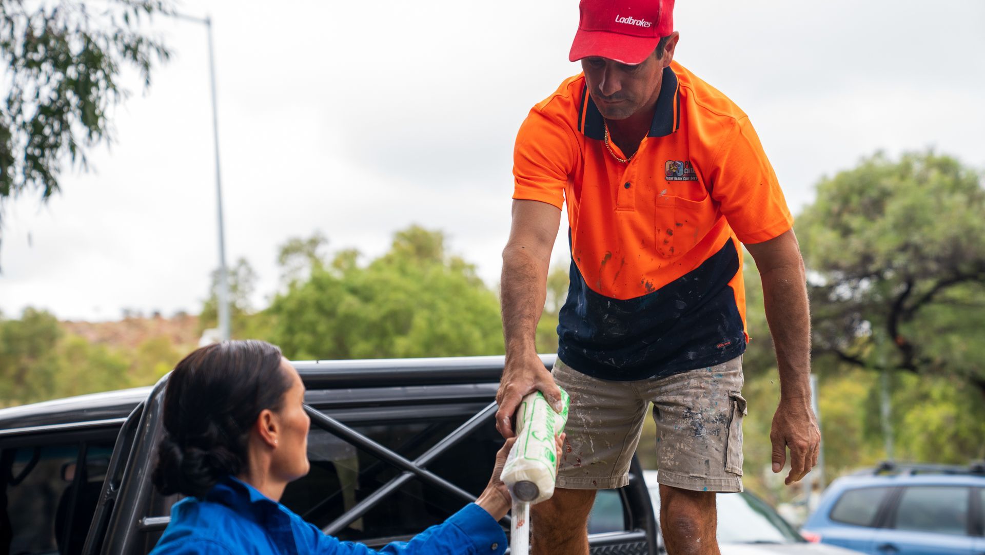 A man in an orange shirt is giving a woman a roll of toilet paper.