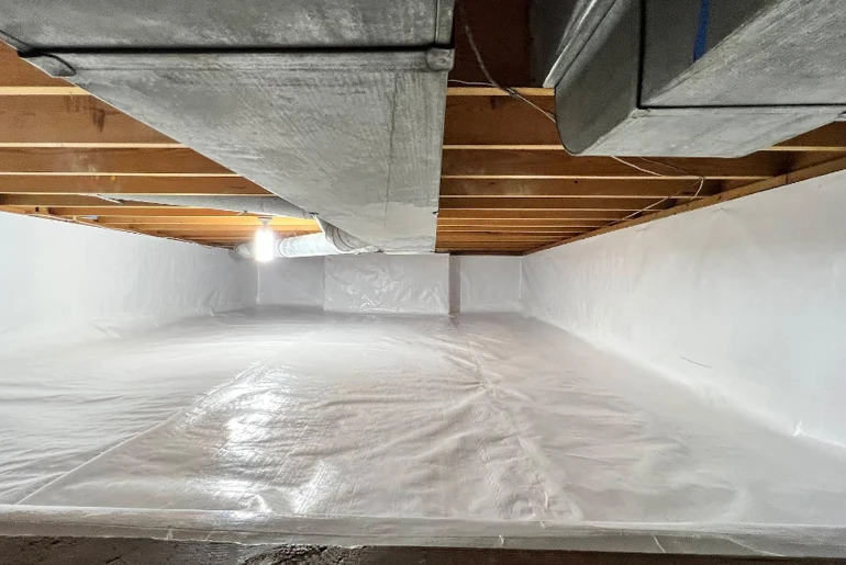 A crawl space encapsulated with white vapor barrier plastic on the floor and walls under wooden joists and ductwork.