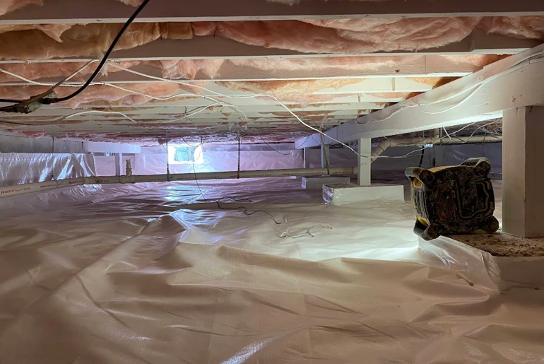 A clean crawl space with a white vapor barrier covering the ground, insulation between joists, and a dehumidifier.