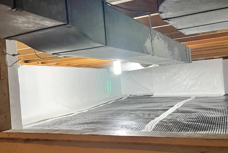 A crawl space featuring a white vapor barrier on the walls, a dimpled plastic floor liner, and a large metal HVAC duct.