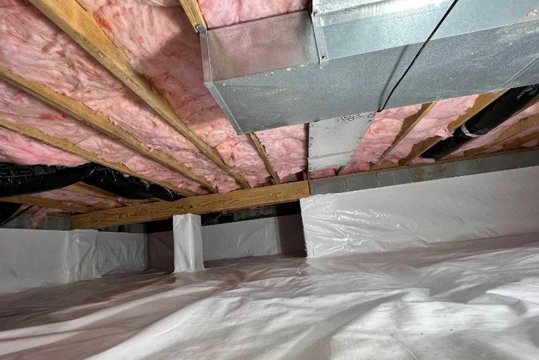 A crawl space with pink fiberglass insulation between wooden floor joists, metal ductwork, and a white vapor barrier floor.