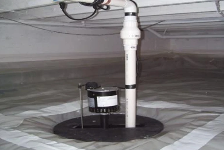 A sump pump installed in a crawl space with a white PVC discharge pipe and a grey vapor barrier on the floor.
