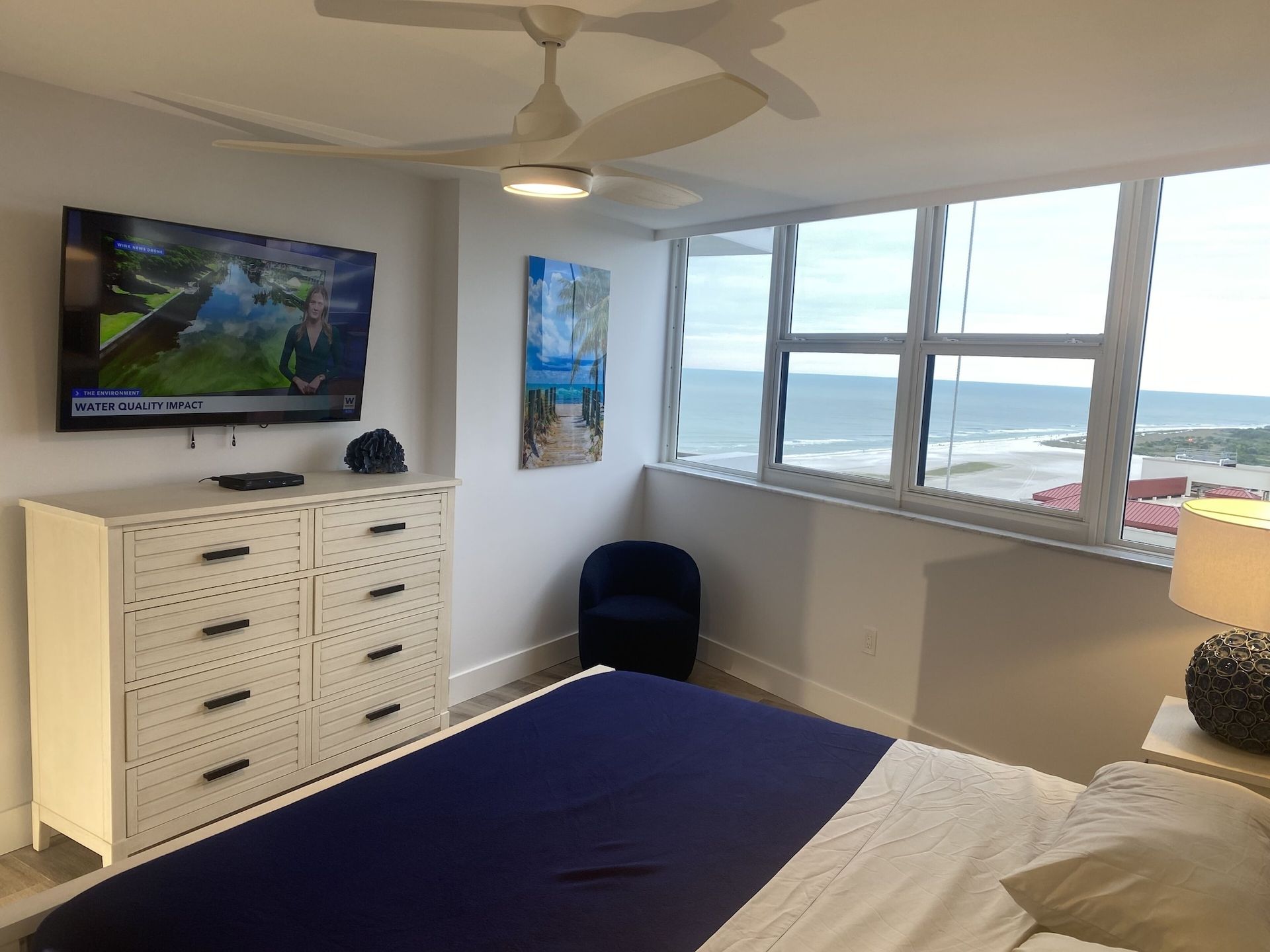 A bedroom with a bed , dresser , television and a view of the ocean.