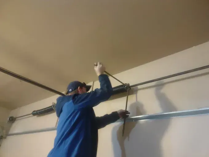 Man in blue jacket repairs a garage door spring while standing on a ladder.