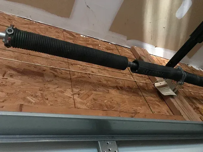 Garage door spring with black protective cover, mounted on a wooden structure above the door.