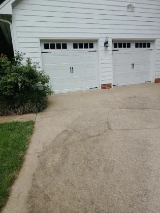White garage doors on a driveway with visible cracks and a bush on the side.