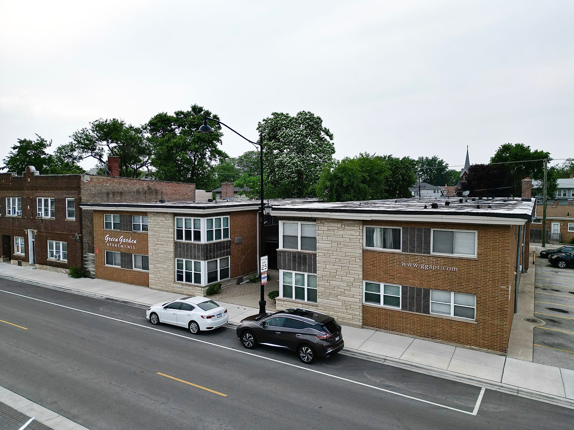 Two brick apartment buildings, parked cars on street. Trees and overcast sky in the background.