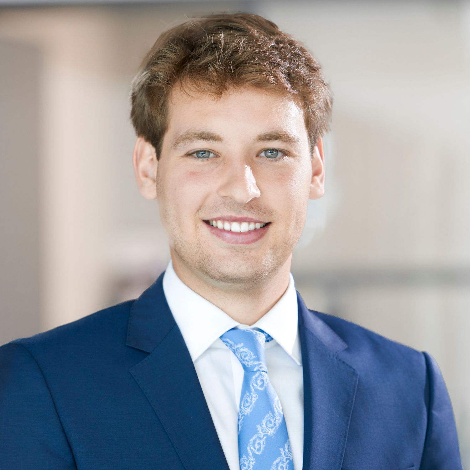 Man with glasses and beard, wearing a gray blazer over a plaid shirt, smiling in front of a white wall.