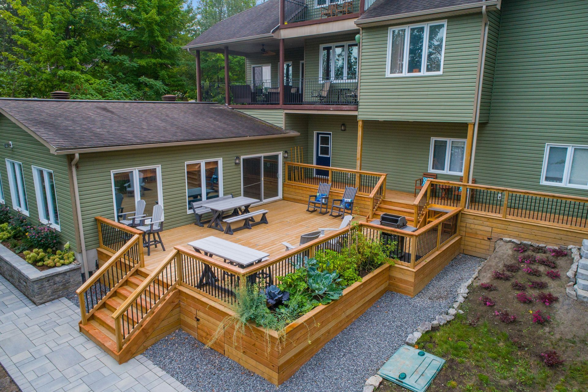 Wooden deck with stairs and seating beside a green house, surrounded by garden beds.