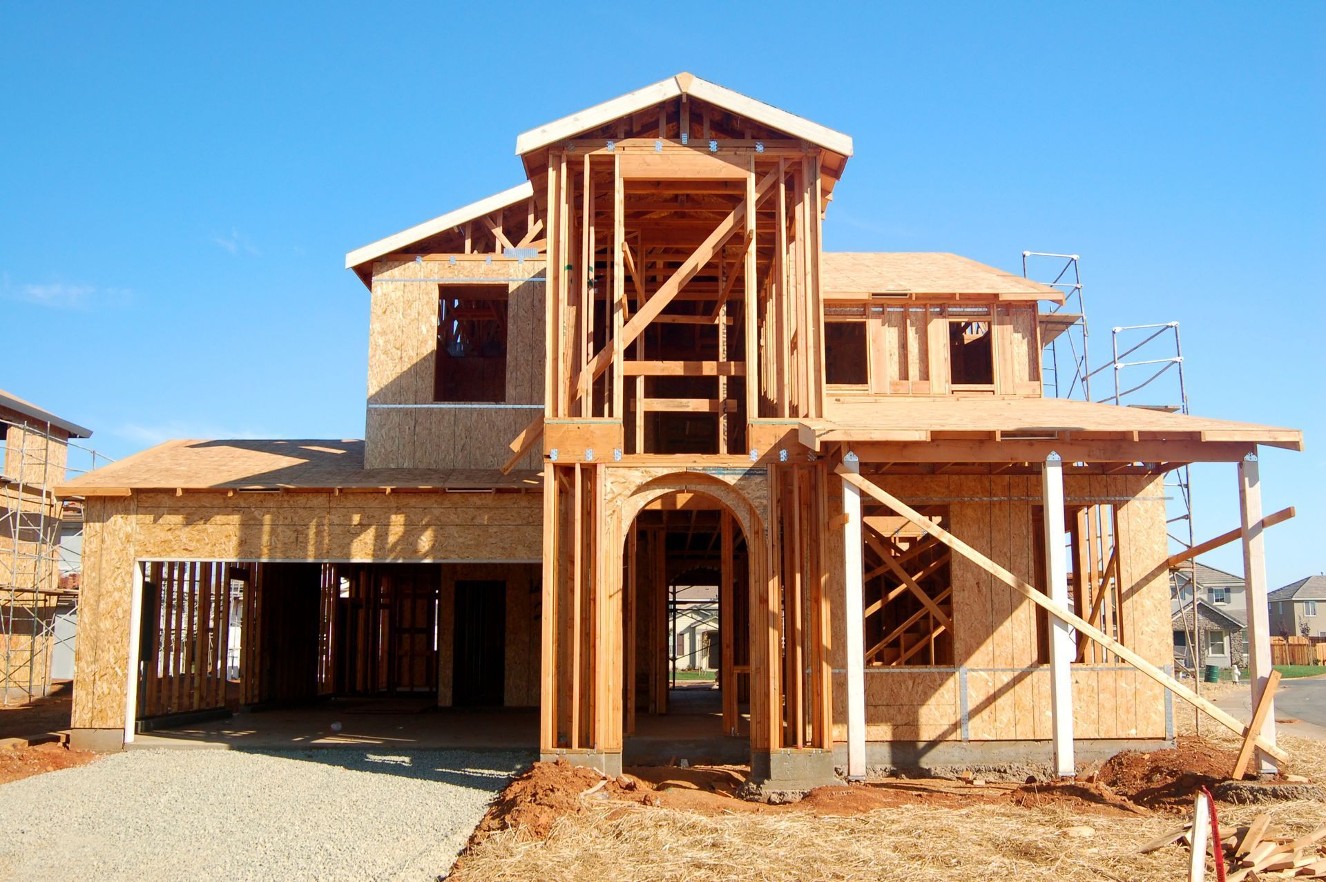 Une maison à ossature bois de deux étages en construction, se détachant sur un ciel bleu azur, avec une entrée voûtée et un garage ouvert.