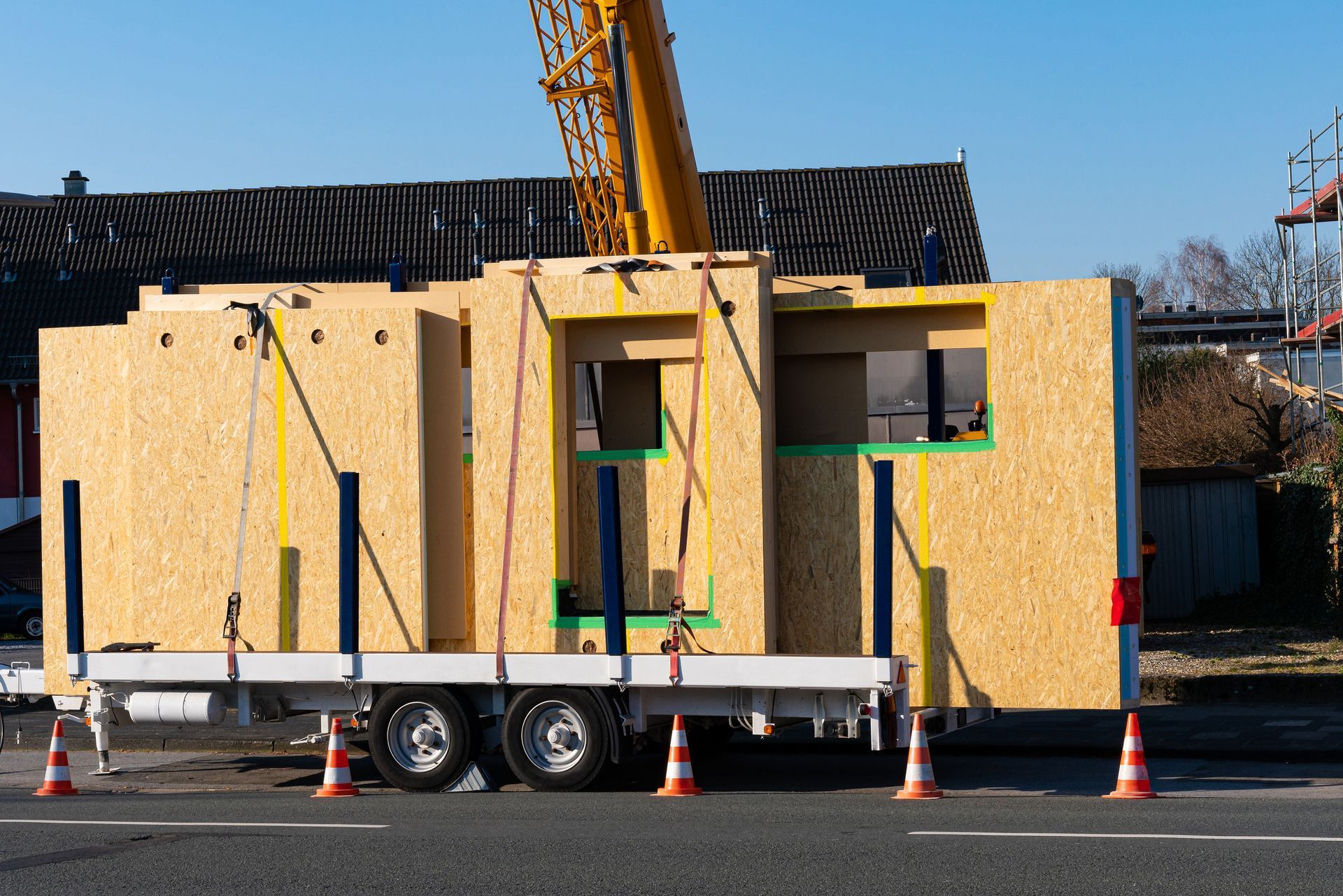 Une grue soulève un élément de mur préfabriqué en bois d'une maison depuis une remorque à plateau, dans une rue bordée de cônes de signalisation orange.