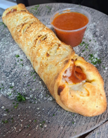 A pepperoni roll is sitting on a wooden table next to a cup of sauce.