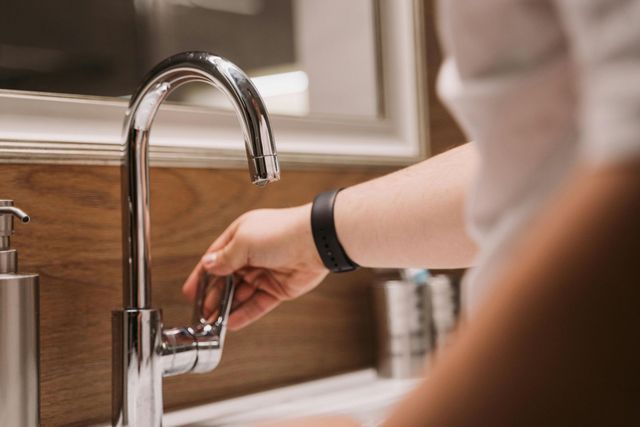 Person's hand turning on a chrome faucet in a bathroom, near a soap dispenser and against a wood panel wall.