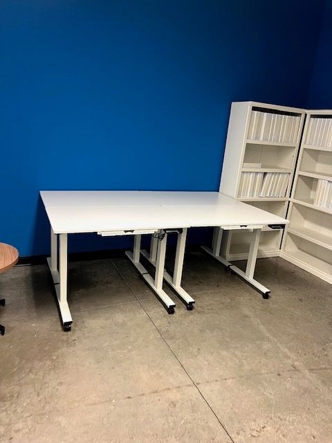 White desk against blue wall with a white shelf unit. Desk has black legs, and is on wheels.