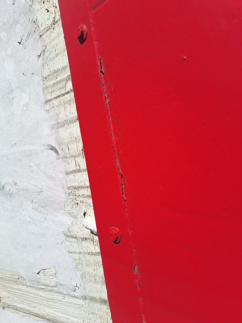 Red painted metal panel attached to a textured, light-colored surface with rivets.