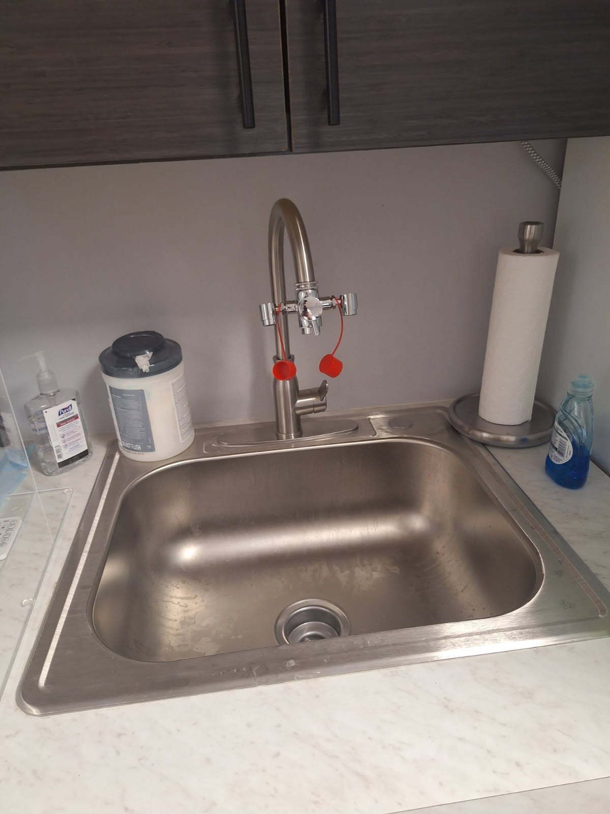Stainless steel kitchen sink with faucet, paper towel holder, and hand sanitizer.