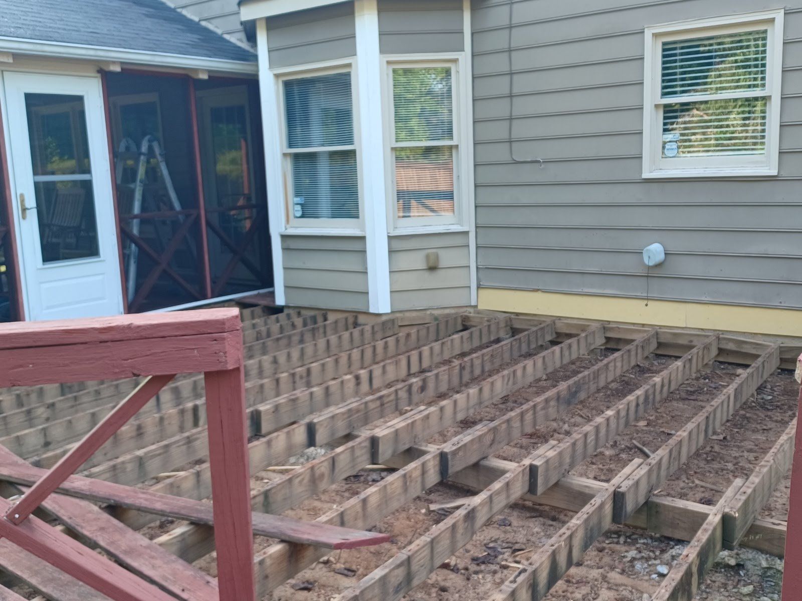 Deck with removed planks, exposing supporting structure. House exterior with windows and door in background.