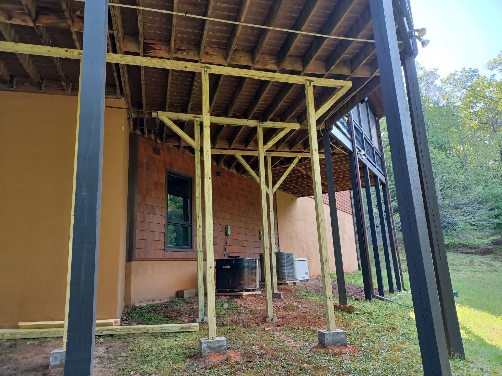 Deck supported by light yellow beams and dark columns, next to a brick building, and lush green grass.