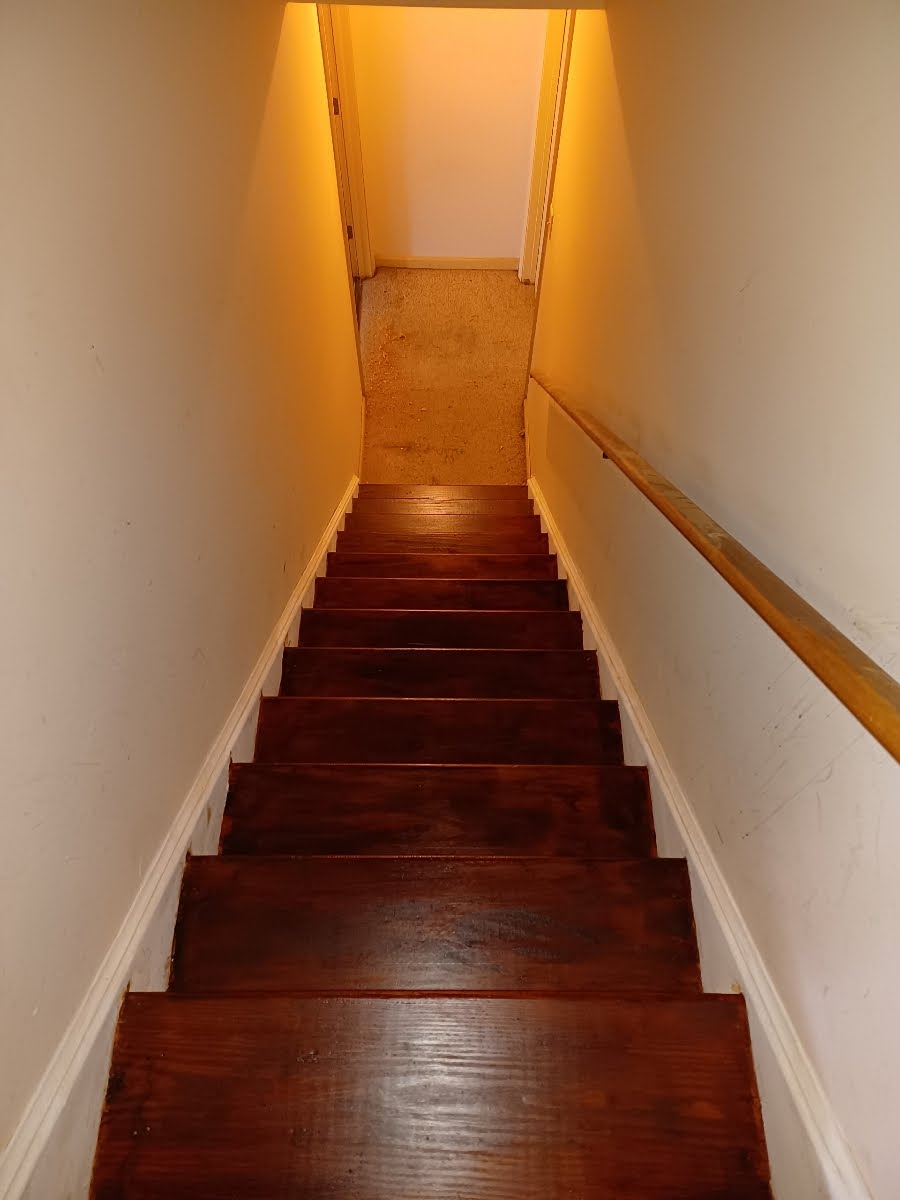 Wooden staircase going upwards, with a handrail on the right and white walls.