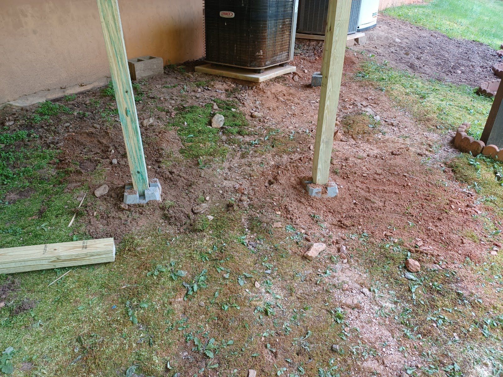 Two wooden deck posts on concrete blocks, set in dirt and grass near an AC unit.