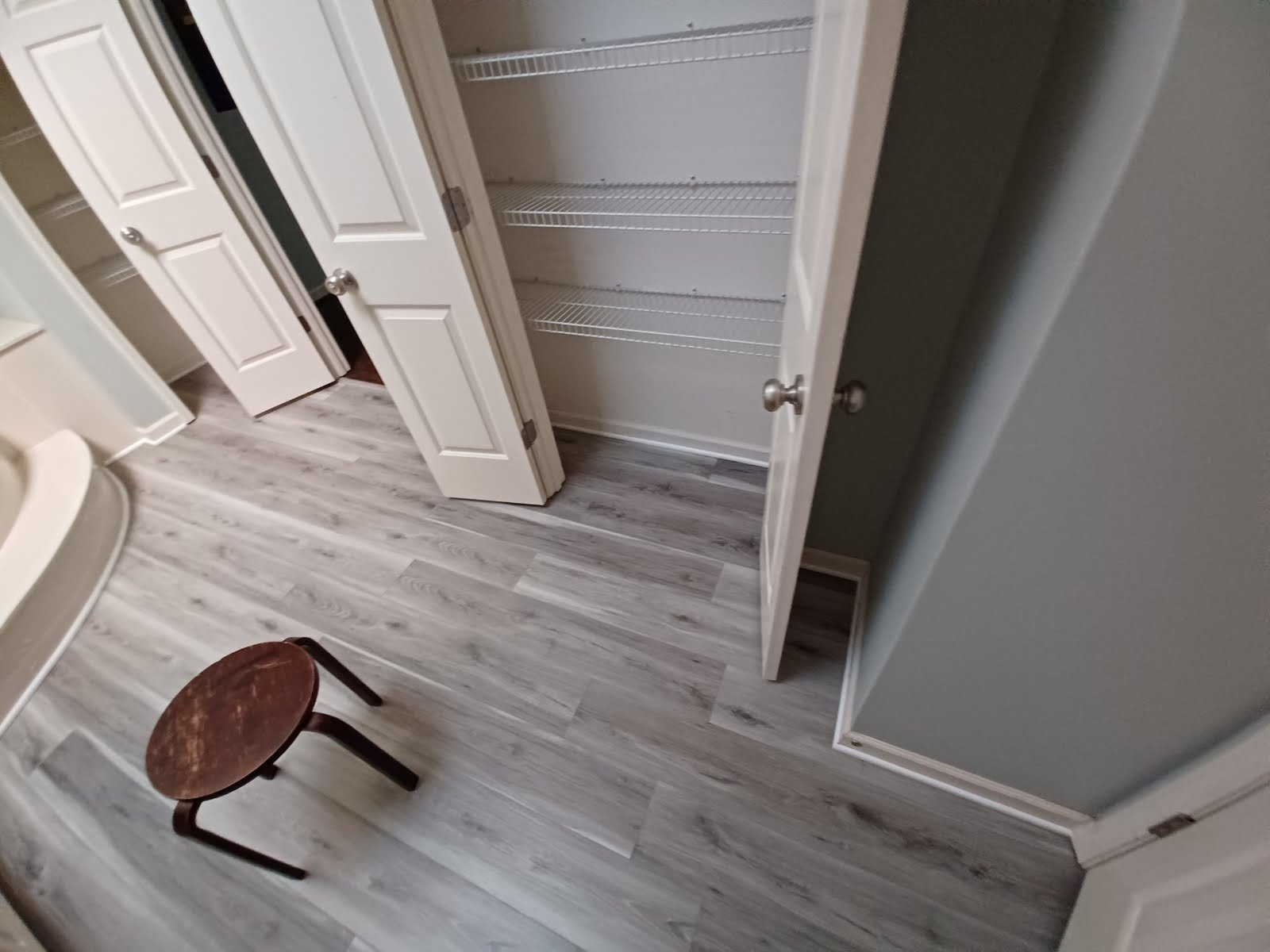 Open closet with wire shelving, white doors, and wooden stool on gray-tone wood flooring.