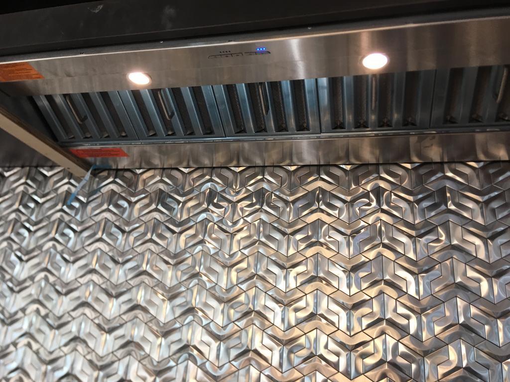 Stainless steel range hood above textured silver tile backsplash.
