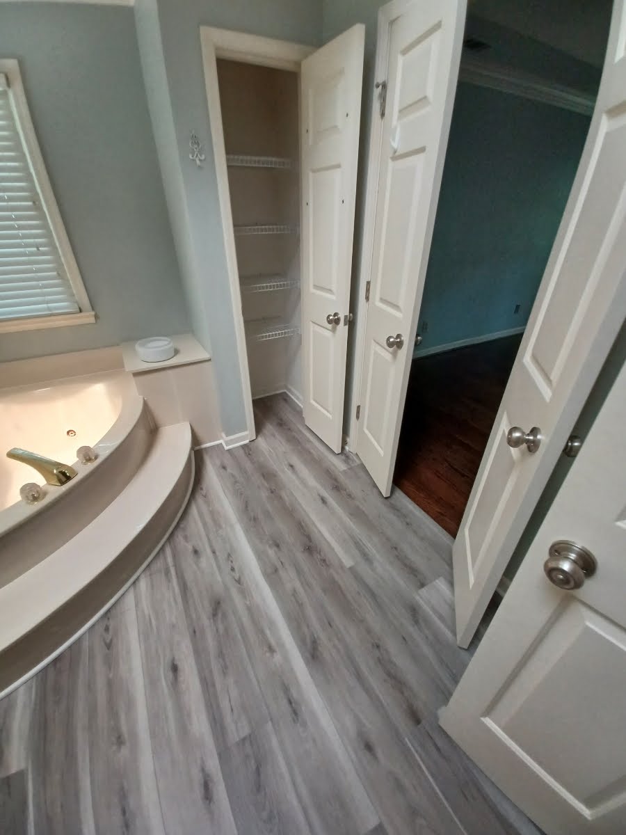 Bathroom with gray wood-look flooring, light blue walls, a bathtub, and open doors to a closet and a room with wood floors.
