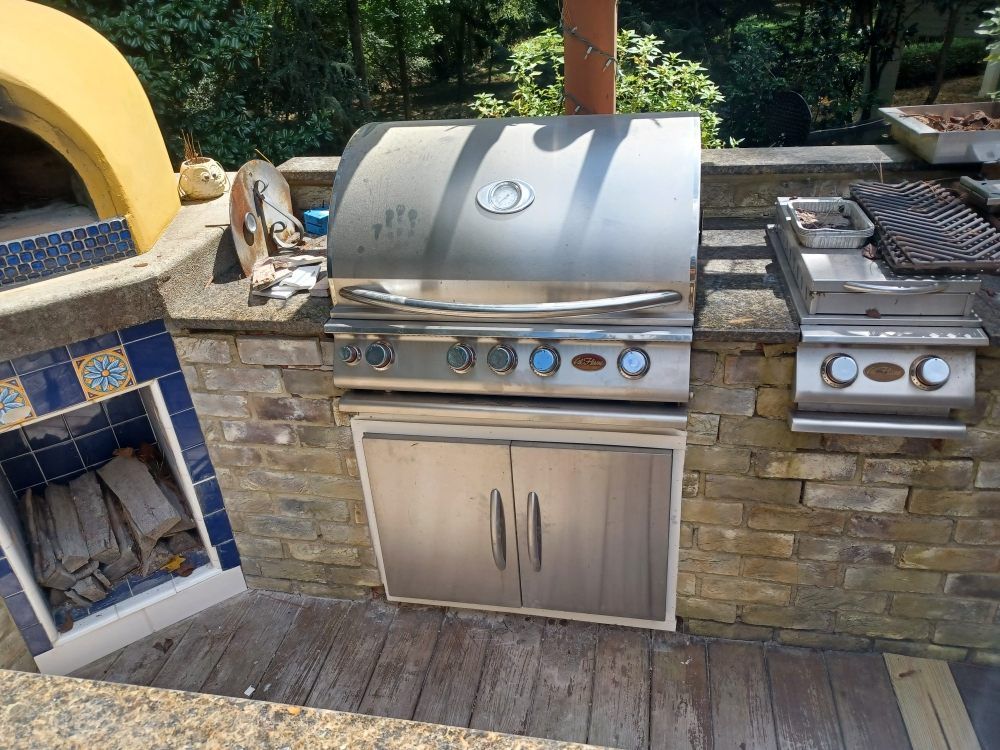 Stainless steel outdoor grill and side burner built into a stone and brick counter with a wood deck and pizza oven.