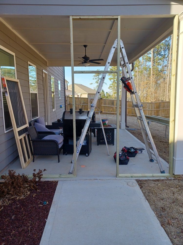 Porch screening project in progress: Wooden frame, ladder, tools, patio furniture, concrete floor, sunny day.