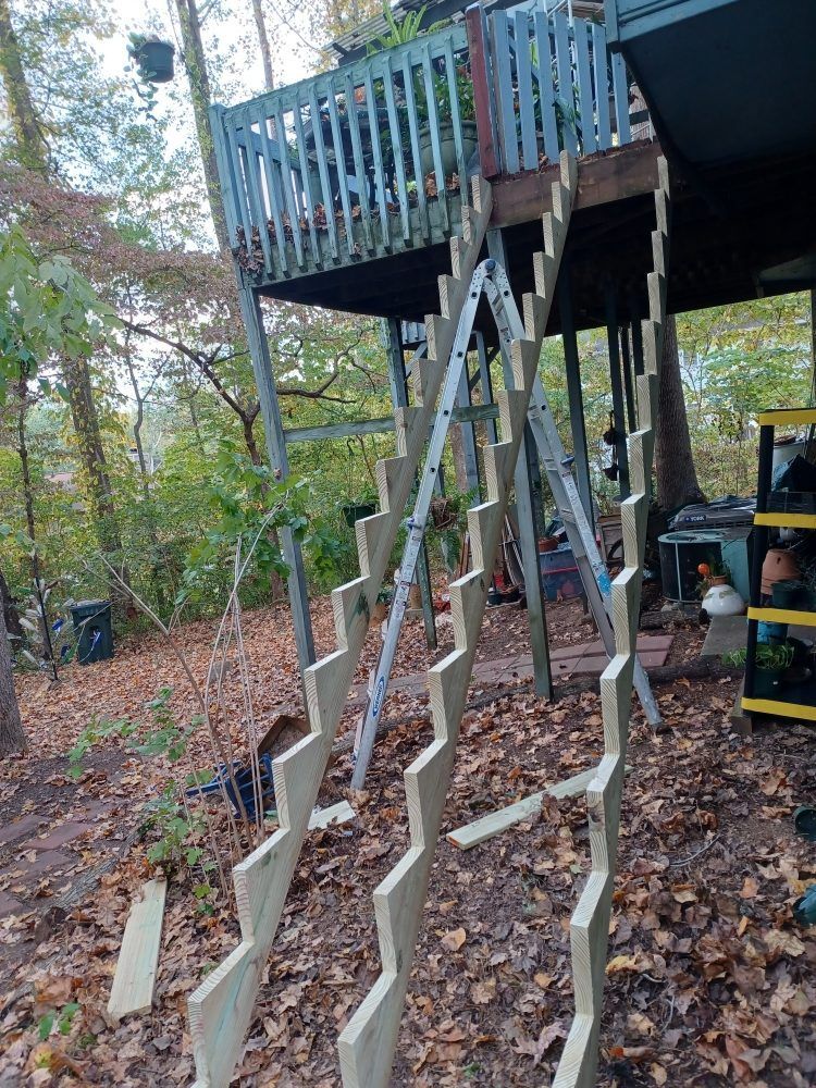 Wooden stairs under construction leading to a deck, supported by a ladder and wooden framework. Outdoors with trees.