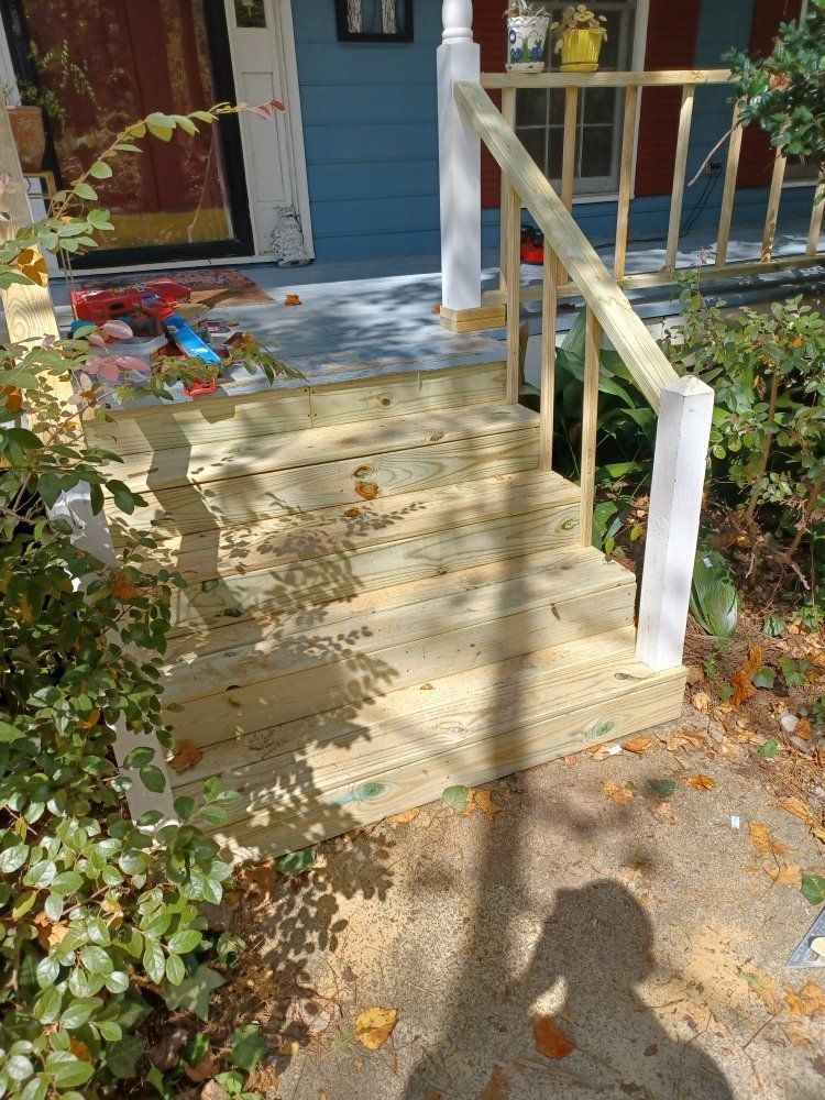 Wooden front porch steps with white railings lead to a blue house, surrounded by foliage.