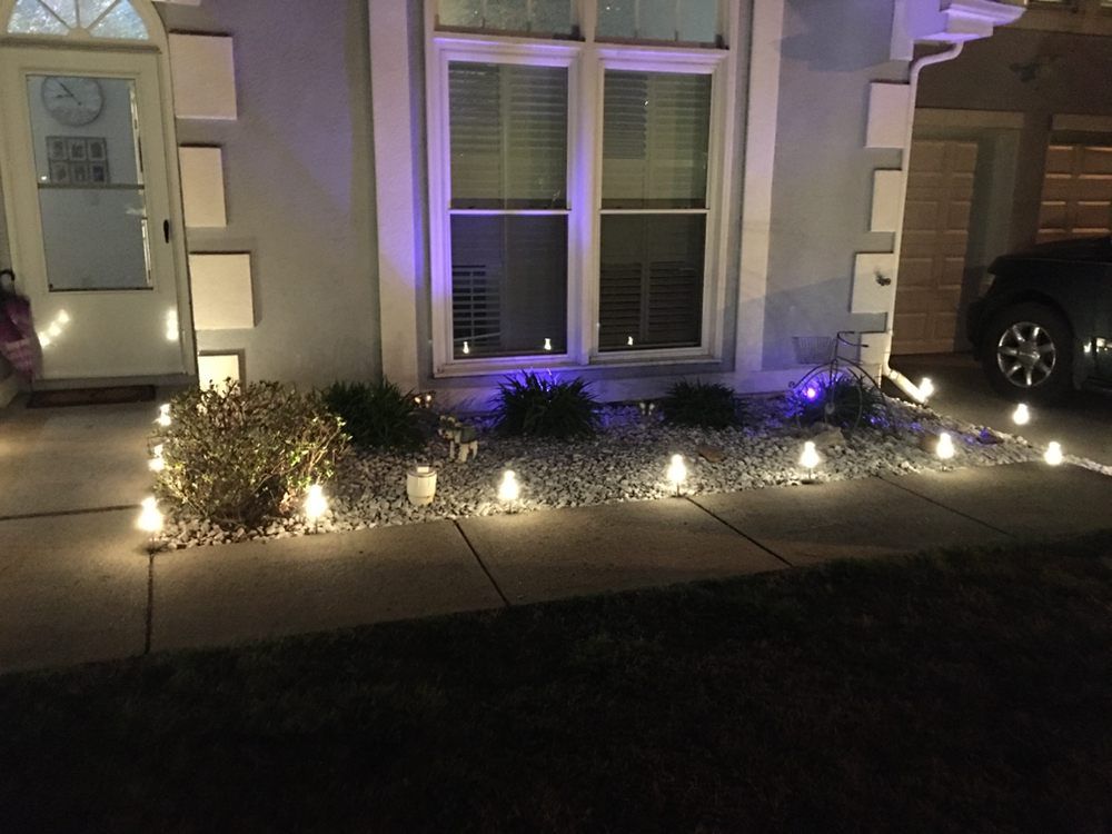 Path lights illuminating a flower bed in front of a house at night; a car is parked in a driveway.