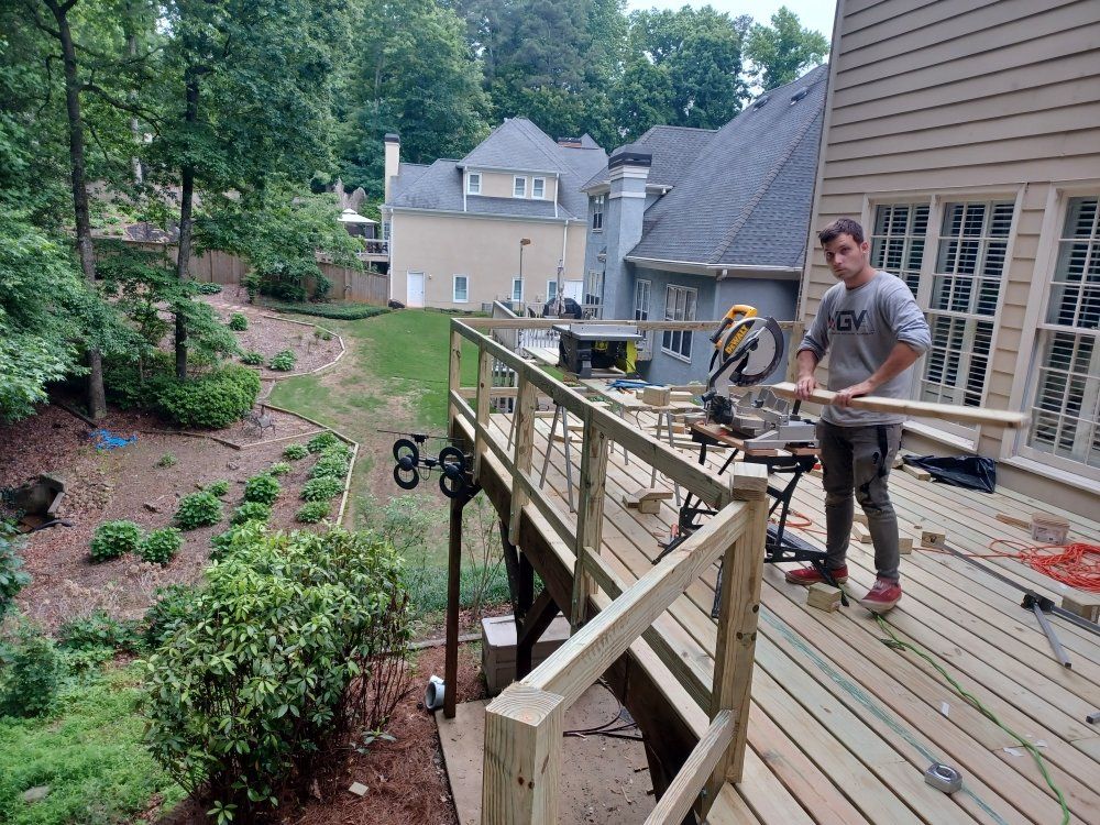 Man on a deck, using a saw to cut wood. Houses and trees are visible in the background.