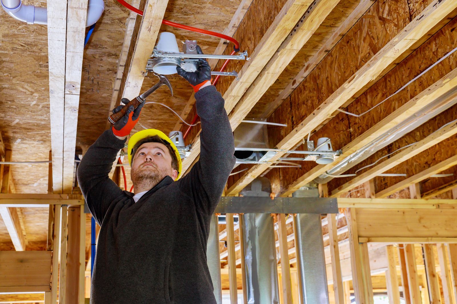 Construction worker in hard hat, installing electrical fixtures in a wooden ceiling frame.